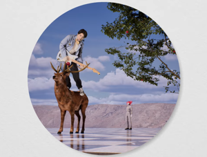 a circular framed shot of a guy on top of a deer with a distant teto in the backdrop who is standing under the branches of a tree. a distant mountain range spans the backdrop.